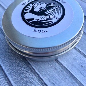 May include: A silver tin with a white label that reads "Manly Hand Healer" and "2 oz." The label features a black and white illustration of a welder wearing a welding mask and welding a piece of metal.