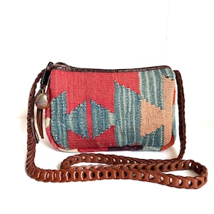 Puede incluir: Un bolso rectangular tejido con una correa de cuero marrón. El bolso presenta un patrón geométrico rojo, azul y beige. Un pequeño dije plateado y borlas de cuero cuelgan del costado. La correa es de diseño trenzado.