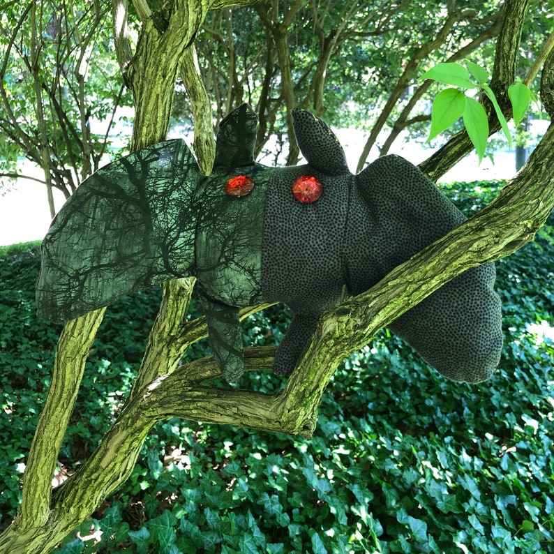 Peut inclure: Un jouet en peluche en forme de cr&eacute;ature avec un motif de camouflage et un corps noir. Le jouet a des yeux en boutons rouges et est perch&eacute; sur une branche d'arbre.