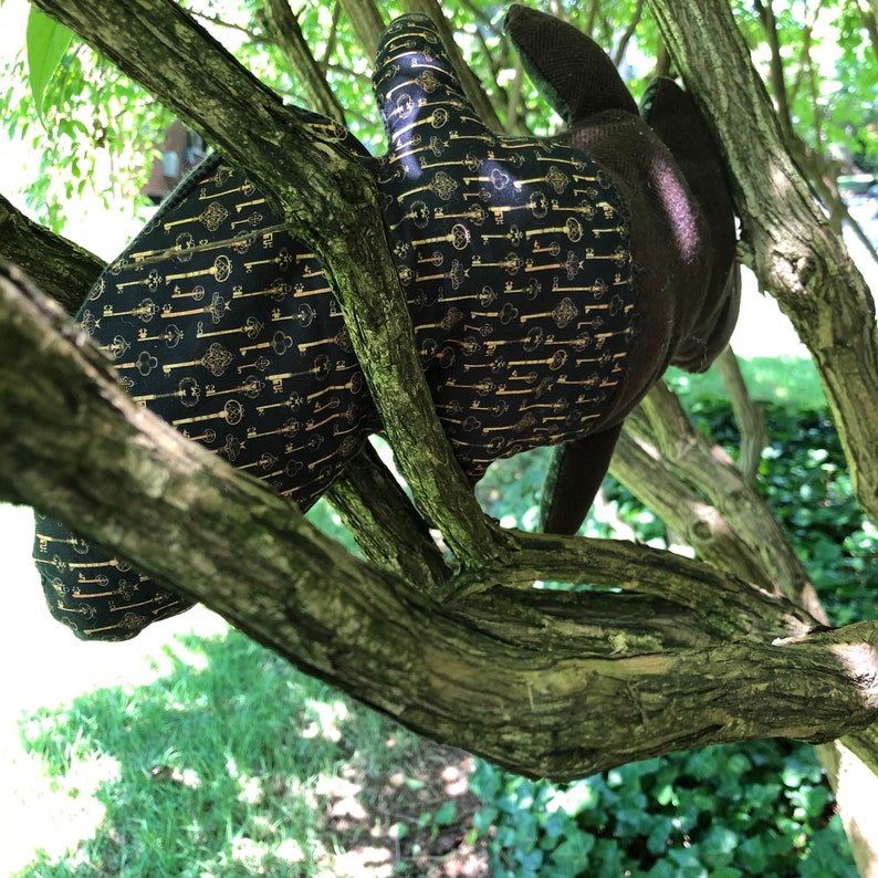 Peut inclure: Un gant en tissu noir avec un motif de cl&eacute;s dor&eacute;es est coinc&eacute; dans une branche d'arbre. Le gant est fait d'un tissu doux et pelucheux et a une surface textur&eacute;e.