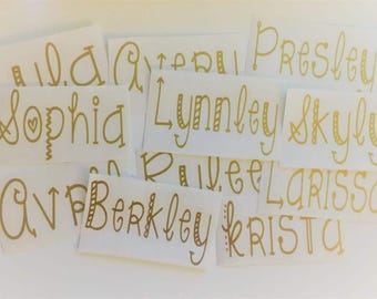 May include: A collection of white rectangular labels with gold-colored handwritten names, including "Sophia", "Lynley", and "Skylin". The names are in a decorative script font, with some names featuring heart and dot embellishments.