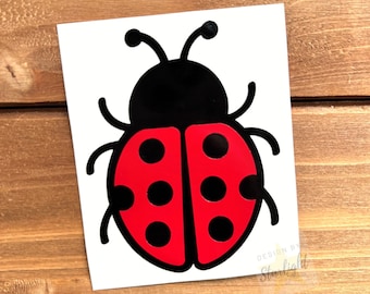 May include: A white sticker featuring a stylized ladybug design. The ladybug has a black head and body, with red wings adorned with black spots. The sticker is on a wooden surface. The text "DESIGN BY Starlight" is in the bottom right corner.