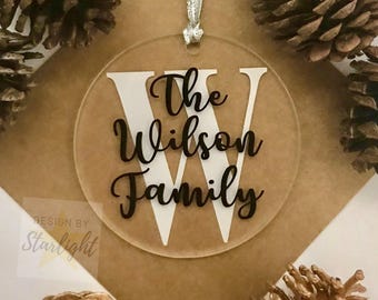 May include: A clear round acrylic bauble with the text "The Wilson Family" and a white letter "W" in the centre. The bauble is hanging from a silver string.
