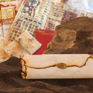 以下が含まれることがあります： 金色の封印と焦げた縁のデザインが施されたヴィンテージスタイルの封筒。 アンティークな地図と赤いカードを背景に、質感のある茶色の表面に置かれています。 テーマは歴史的でヴィンテージです。