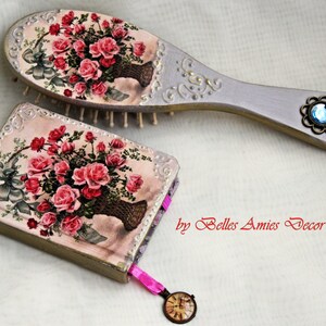 Puede incluir: Un conjunto de dos artículos decorativos con un diseño floral vintage con rosas rosas. El conjunto incluye un cepillo para el cabello con un mango gris claro y un pequeño libro con una cubierta rosa y dorada. El libro tiene un marcapáginas de cinta rosa y un pequeño encanto de reloj.
