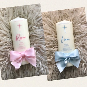 May include: Two white baptism candles with a cross and a name on each. The candle on the left has a pink bow and the text "The Baptism of Rosie Joan Mullan Baptised on 19 March 2023 at St Mary's Church". The candle on the right has a blue bow and the text "The Christening of Liam Sean Carter Christened on 19 March 2023 at St Anthony's Church".