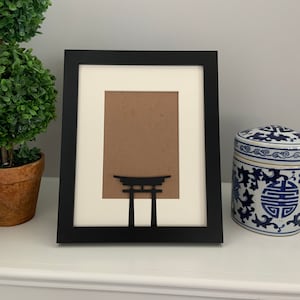 May include: Black picture frame with a silhouette of a Japanese torii gate. The frame is on a white surface with a blue and white porcelain jar in the background.