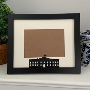 May include: Black picture frame with a white mat and a silhouette of the White House in the center. The frame is on a white surface.
