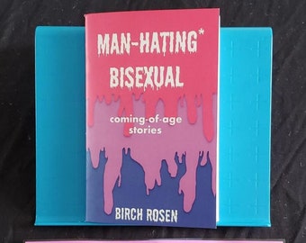 以下が含まれることがあります： ピンクと青の表紙の「MAN-HATING* BISEXUAL」というタイトルの本が、青いブックスタンドに立てられています。本のすぐ下には、「SICK SAD TRANS FAGS」という言葉がコラージュ風に書かれたピンク色のステッカーがあります。背景は黒です。