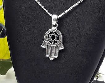 May include: A silver necklace with a hamsa hand pendant featuring a Star of David inside. The necklace is displayed on a black jewelry stand.