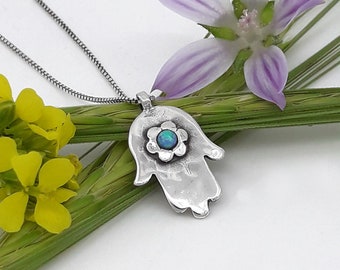 May include: A silver Hamsa hand charm necklace with a blue opal flower in the center. The necklace is on a silver chain and is displayed on a green stem with yellow flowers.