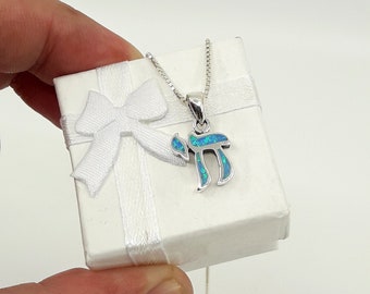 May include: A silver necklace with a blue opal pendant in the shape of the Hebrew letter "Chai". The necklace is in a white gift box with a white ribbon.