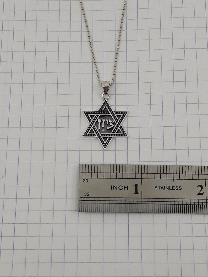 Puede incluir: Un collar de cadena de plata con un colgante de plata en forma de Estrella de David que presenta la palabra hebrea "Chai" en el centro. El colgante tiene un di&aacute;metro de aproximadamente 2,5 cm.