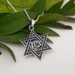 Puede incluir: Un collar de cadena de plata con un colgante de estrella de David de plata. El colgante tiene un acabado de esmalte negro y la palabra hebrea "Chai" inscrita en &eacute;l.