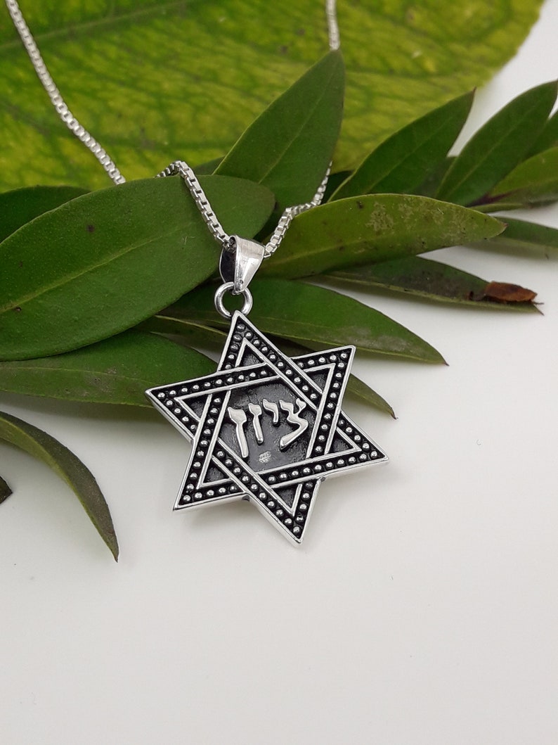 Puede incluir: Un collar de cadena de plata con un colgante de estrella de David de plata. El colgante tiene un acabado de esmalte negro y la palabra hebrea "Chai" inscrita en &eacute;l.