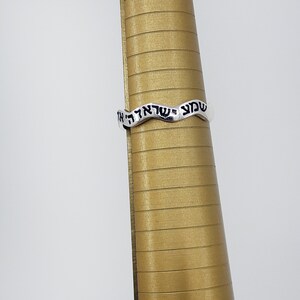 May include: A silver ring with a wavy design and Hebrew text inscribed on it. The ring is on a gold ring sizer.