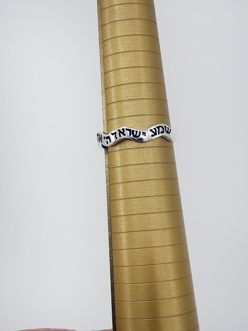 May include: A silver ring with a wavy design and Hebrew text inscribed on it. The ring is on a gold ring sizer.