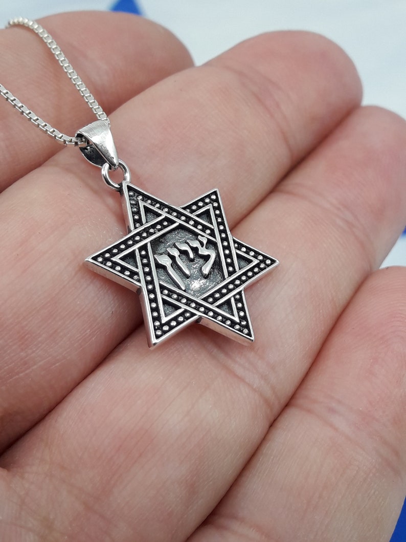 Puede incluir: Un collar con un colgante de plata en forma de Estrella de David con una inscripci&oacute;n hebrea dentro de la estrella. El colgante est&aacute; en una cadena de plata.