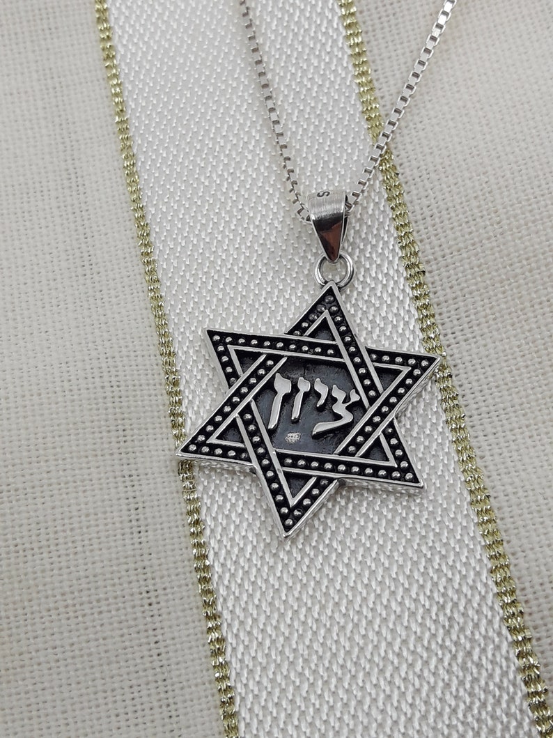 Puede incluir: Un collar de cadena de plata con un colgante de estrella de David de plata. El colgante tiene un acabado de esmalte negro y la palabra hebrea "Chai" inscrita en el centro.