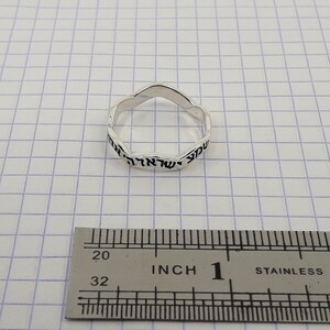 May include: A silver ring with Hebrew writing around the band. The ring is measured against a ruler, showing it is 1 inch in diameter.