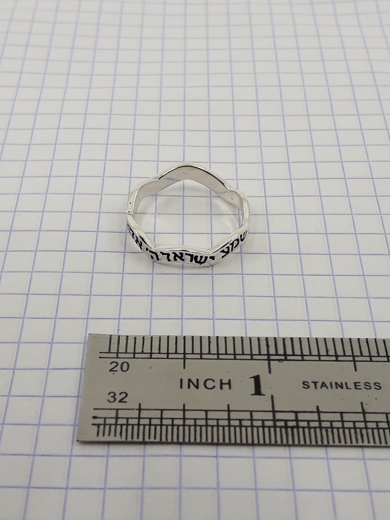 May include: A silver ring with Hebrew writing around the band. The ring is measured against a ruler, showing it is 1 inch in diameter.