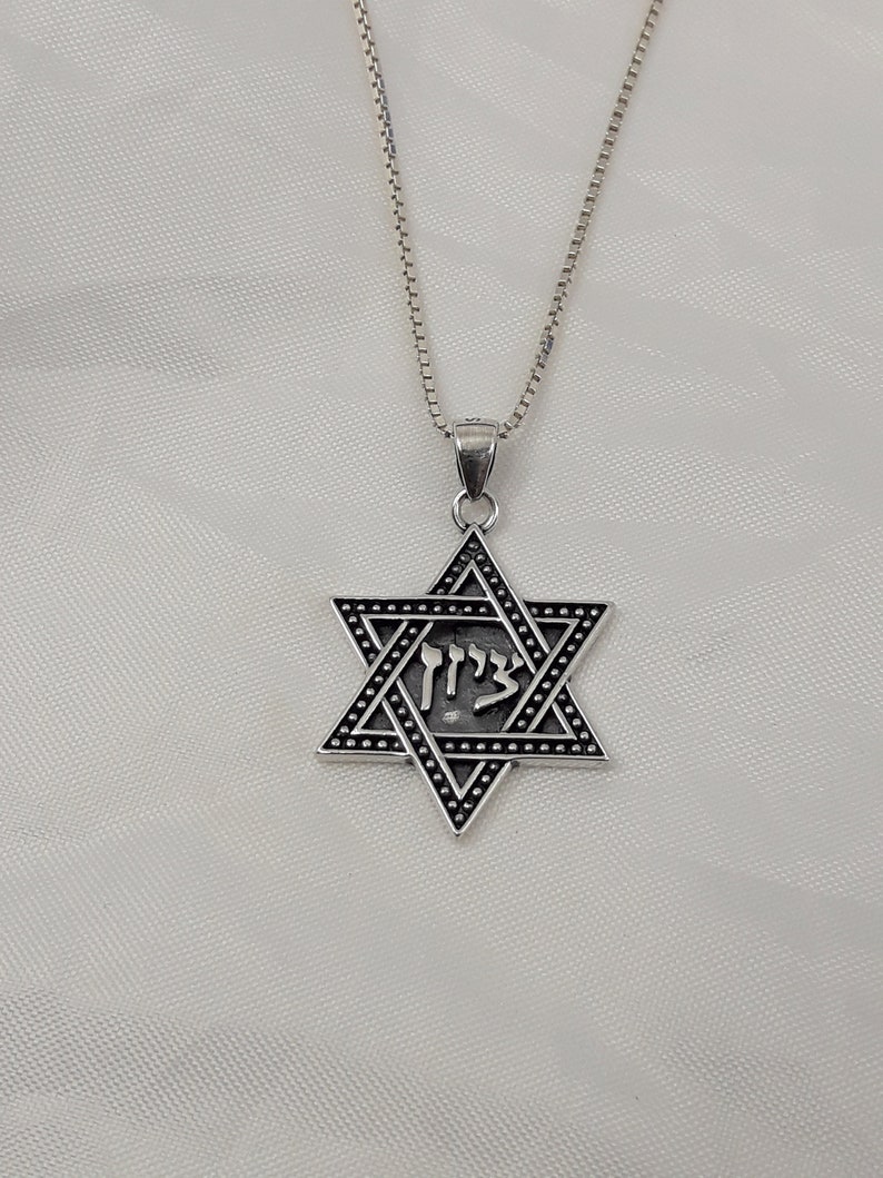 Puede incluir: Un collar de cadena de plata con un colgante de plata en forma de estrella de seis puntas con la palabra hebrea "Chai" inscrita en su interior.