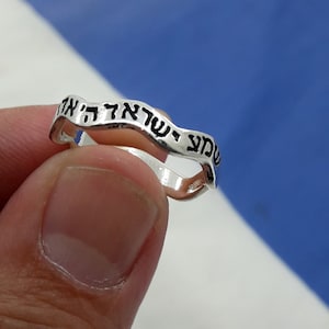 May include: A silver ring with a wavy band inscribed with Hebrew text. The text reads "Shema Yisrael Adonai Eloheinu Adonai Echad".