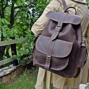 May include: A brown leather backpack with a flap closure and multiple pockets. The backpack has a distressed look and is made of high-quality leather.