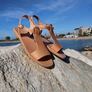 Peut inclure: Une paire de sandales en cuir marron clair avec une bride en T. Les sandales sont sur un rocher avec une &eacute;tendue d'eau bleue en arri&egrave;re-plan.