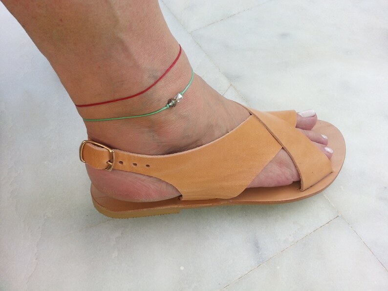 Puede incluir: Sandalias de cuero marr&oacute;n claro con cierre de hebilla y dise&ntilde;o de tiras cruzadas. Las sandalias se llevan en un pie con dos tobilleras, una roja y una verde.
