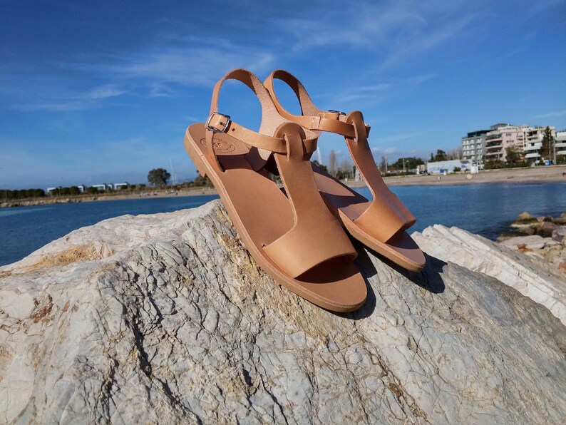 Peut inclure: Une paire de sandales en cuir marron clair avec une bride en T. Les sandales sont sur un rocher avec une &eacute;tendue d'eau bleue en arri&egrave;re-plan.