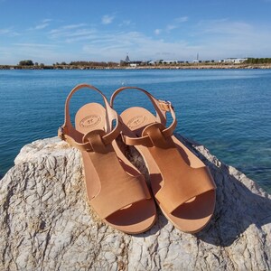 Peut inclure: Une paire de sandales en cuir tann&eacute; avec une bride en T, pos&eacute;es sur un rocher avec une &eacute;tendue d'eau bleue en arri&egrave;re-plan.