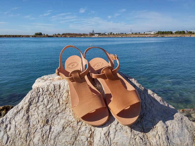Peut inclure: Une paire de sandales en cuir tann&eacute; avec une bride en T, pos&eacute;es sur un rocher avec une &eacute;tendue d'eau bleue en arri&egrave;re-plan.