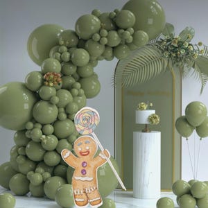 May include: A green balloon arch with a gingerbread man cutout holding a lollipop. The arch is decorated with green leaves and a cake on a white pedestal. The text "Happy Birthday" is written on a green background.