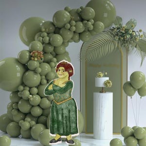 Pode incluir: Um arco de balões verdes com um fundo verde e dourado. Um personagem com um vestido verde e uma coroa está em frente ao arco. Um bolo branco com flores verdes está em cima de um pedestal branco na frente do fundo. O fundo tem o texto "Happy Birthday" escrito nele.