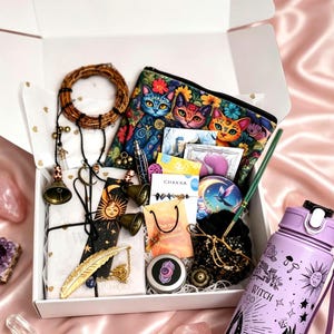 May include: A white gift box containing a zippered pouch with a colourful cat print, a light purple water bottle with black celestial graphics and the word WITCH, a gold feather bookmark, small bells, a sun and moon bookmark, and various crystals.