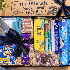 May include: A black gift box containing a blue raspberry sweet packet, a Cadbury Dairy Milk chocolate bar, a yellow butter-themed box, a blue football patterned pencil case, pens, a book review notepad, and a stack of books sticker, all nestled in shredded paper.