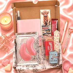 May include: A cardboard gift box containing a pink notebook, a Highland cow bookmark, a chocolate raspberry coffee packet, pink glittery under-eye patches, a clear speckled hair clip, a Lotus Biscoff snack, a floral nail file, and a Book Worm keychain.