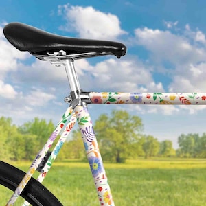 Peut inclure: Un cadre de vélo blanc avec un motif floral dans des tons de rose, violet, jaune et bleu. Le vélo a une selle en cuir noir.