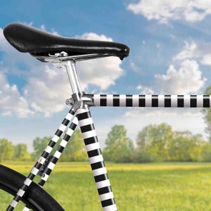 Könnte beinhalten: Ein schwarz-weiß gestreiftes Fahrradrahmen mit einem schwarzen Ledersattel. Das Fahrrad steht auf einem Feld mit grünem Gras und blauem Himmel im Hintergrund.