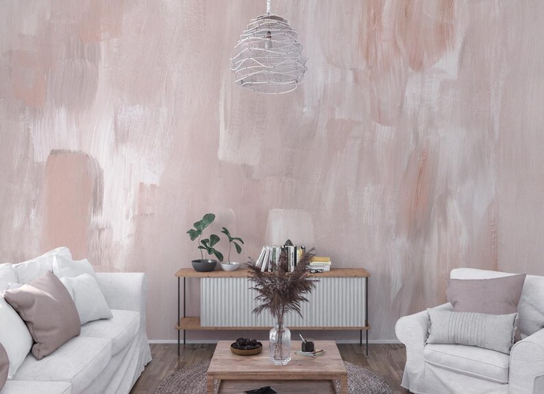 May include: A living room featuring a white sofa and a white armchair. The room has a light pink wall with white brushstrokes. A white wire cage light fixture hangs from the ceiling. A wooden coffee table with a vase of dried flowers sits in front of the sofa and armchair.