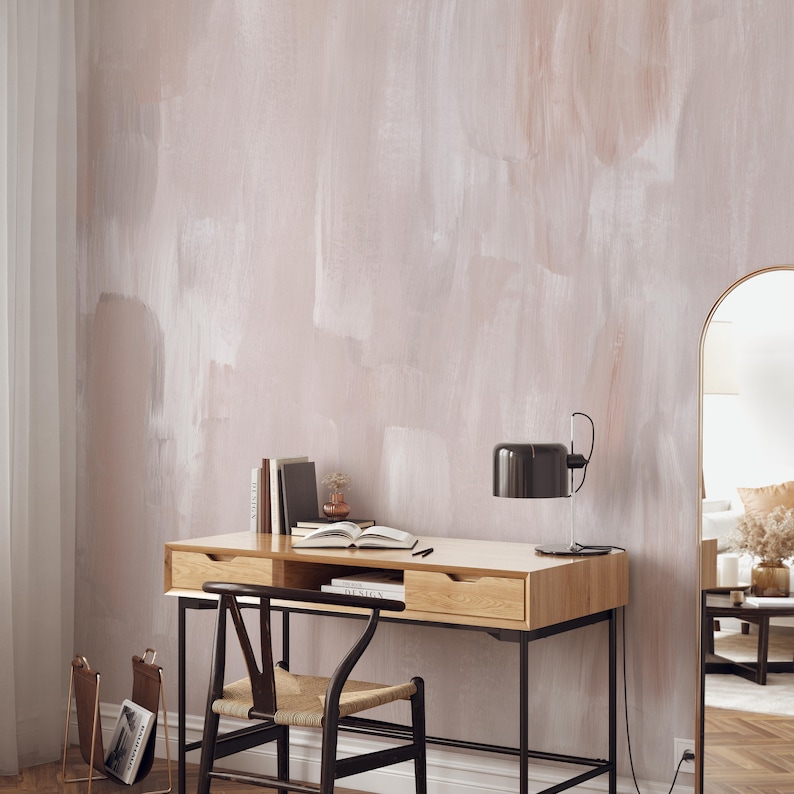 May include: A light wood desk with two drawers and black metal legs. A black lamp with a white shade sits on the desk. A chair with a woven seat and black metal legs sits in front of the desk. The desk is in front of a wall with a light pink and white abstract paint design.