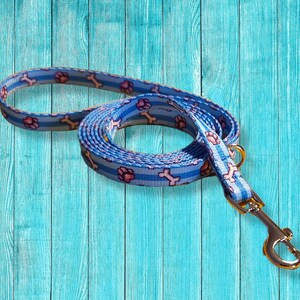 May include: A blue and white dog leash with a pattern of pink paw prints and bones. The leash has a metal clasp and a loop for the owner's hand. The background is a light blue wooden surface.