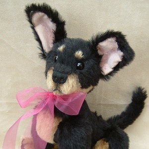 May include: A black and tan plush toy dog with a pink ribbon around its neck. The dog is sitting with its head tilted and looking up.