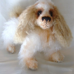 Puede incluir: Un perro de peluche blanco y marrón con pelo largo y esponjoso. El perro tiene ojos marrones y una nariz negra.