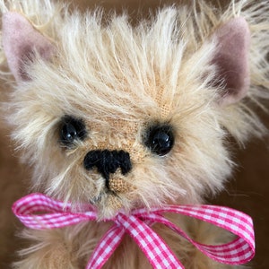 May include: A small, fluffy, light brown teddy bear with pink ears and black eyes. The bear is wearing a pink and white gingham ribbon around its neck.