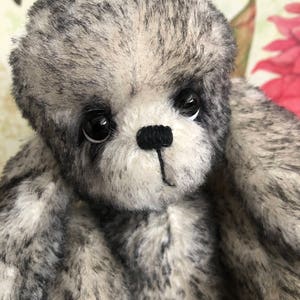 May include: A close-up of a grey and white plush toy bear with black eyes and a black nose. The bear has a soft, fluffy texture.