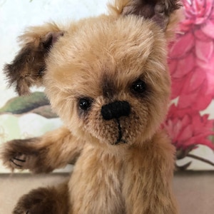 May include: A small, brown teddy bear with a fluffy coat and black eyes. The bear is sitting up with its right arm raised and its left arm resting on its lap.