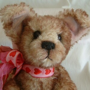 May include: A brown teddy bear with floppy ears wearing a pink ribbon with red hearts. The bear has a black nose and brown eyes.