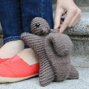 Puede incluir: Un juguete de ardilla de crochet marrón con ojos negros. El juguete está siendo sostenido por una persona que lleva zapatos rojos y pantalones vaqueros azules.